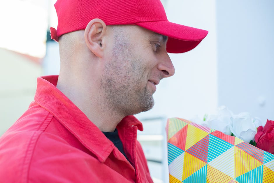 A delivery man wearing a red cap and a red jacket is holding a colorful, geometric-patterned gift bag filled with white and pink flowers. The bouquet features fresh blossoms with visible petals and gr