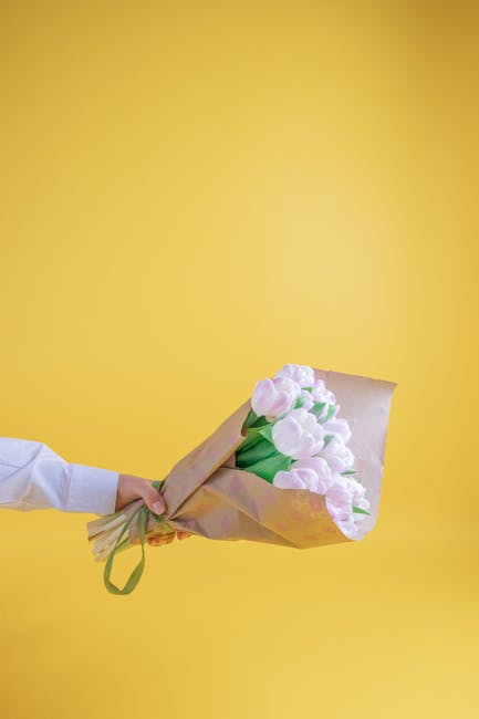 A person wearing a white shirt holds a bouquet of fresh, pastel pink tulips wrapped in light brown paper and secured with a green ribbon. The bouquet is held with one hand, extending horizontally agai