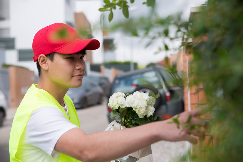 A young delivery person wearing a red cap and neon yellow safety vest is seen outdoors, reaching out to place a fresh floral gift comprising white hydrangeas and roses against a green leafy background