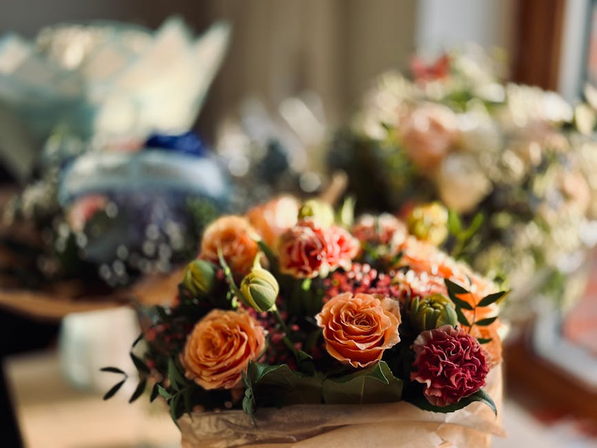 A vibrant bouquet of fresh flowers featuring peach and coral roses, pink carnations, and green foliage arranged in a compact, rounded style. The flowers are wrapped in rustic brown paper, with some gr